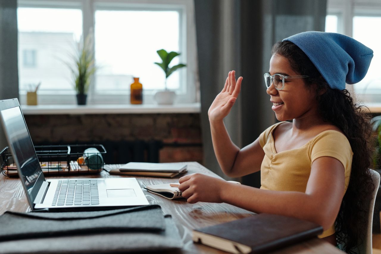A young girl waves at her laptop during a video call in a cozy home office setting.